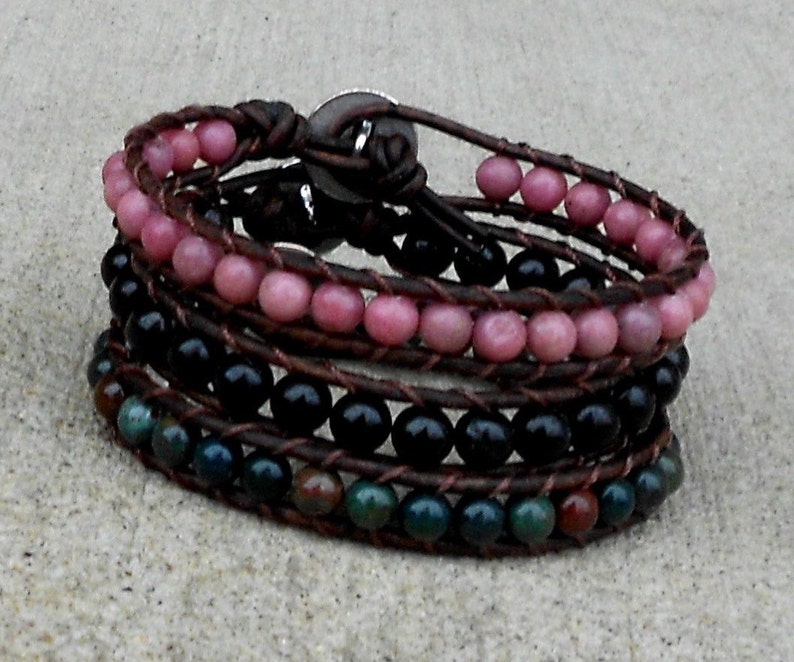 May include: Wrap bracelet featuring multiple strands of beads in pink, black, and green hues. The beads are strung on brown leather cords, creating a layered effect. A button clasp secures the bracelet.