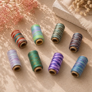 Può includere: Otto rocchetti di filo da ricamo variegato in colori assortiti tra cui rosso, verde, blu, viola e verde acqua. Ogni rocchetto presenta un nucleo in legno e una lucentezza metallica, disposti in modo sparso su una superficie beige testurizzata.
