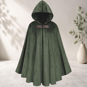Puede incluir: Una capa con capucha de terciopelo verde oliva oscuro que llega hasta el suelo, con una capucha estructurada y un cierre de lengüeta de piel sintética marrón en el cuello con un anillo de metal plateado. La prenda tiene una silueta amplia.
