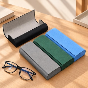 May include: A set of four rectangular eyeglass cases in black, gray, green, and blue, each featuring a smooth exterior and a light gray interior lining. A pair of black-framed eyeglasses with clear lenses is positioned in front of the cases.