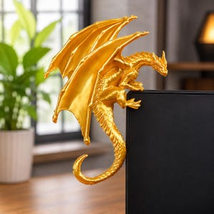 May include: A metallic gold dragon figurine perched on the top corner of a black rectangular monitor. The dragon features detailed scales, large spread wings, and a long, curved tail that hangs down the side of the screen in a resting position.