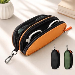 May include: An orange and black zippered pouch with two compartments, containing white-framed glasses. The pouch features a metal carabiner clip attached to the side. Two additional pouches in solid black and olive green are shown separately.