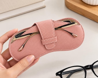 May include: A dusty pink textured eyeglass case with a central strap closure. The case is oval-shaped with visible stitching along the edges. A pair of gold-toned metal frame glasses rests inside the case, and a pair of black frame glasses sits below.