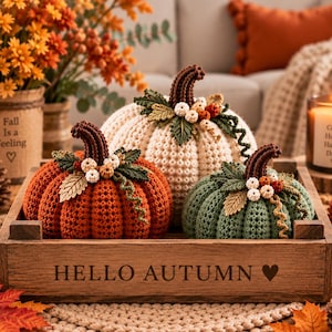 May include: Three crocheted pumpkins in orange, cream, and sage green sit inside a rectangular wooden crate engraved with the text HELLO AUTUMN and a heart. Each pumpkin features a dark brown stem, green leaves, and small white berries.