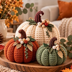 May include: Three crocheted pumpkins in cream, orange, and sage green. Each pumpkin features a dark brown stem, green crocheted leaves, small white and orange berries, and a metallic gold leaf accent. They are arranged together on a round wooden tray.