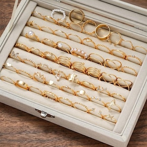 May include: A collection of gold-toned metal rings arranged in a light gray velvet jewelry tray. The rings feature various designs including plain bands, twisted textures, pearl accents, butterfly motifs, and small clear stones in multiple rows.