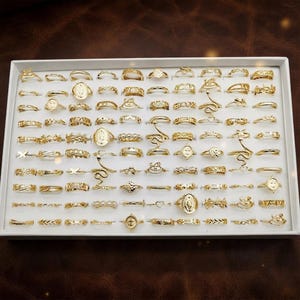 Op de afbeelding: Een wit rechthoekig presenteerblad met een grote collectie goudkleurige metalen ringen. De ringen hebben diverse ontwerpen, waaronder dunne banden, getextureerde patronen, ovaalvormige medaillons, slangenmotieven en hartvormen, gerangschikt in rijen.