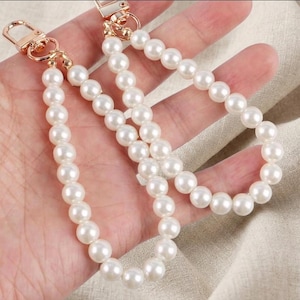 May include: Two decorative bag straps made of round, white faux pearls strung together. Each strap features a polished gold-tone metal lobster clasp at the end, allowing for easy attachment to various accessories or handbags.