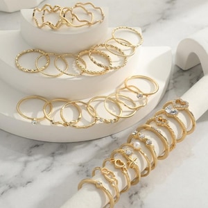 Könnte beinhalten: Eine Sammlung goldfarbener Ringe mit verschiedenen Designs, darunter schlichte Bänder, gedrehte Texturen, wellenförmige Formen sowie Ringe mit kleinen klaren Kristallen, einem herzförmigen Stein und einer einzelnen weißen Perle.