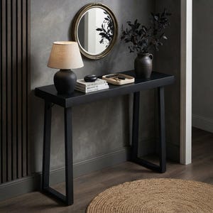 Puede incluir: Una mesa consola negra con tablero rectangular y patas en forma de U. Sobre la mesa hay una lámpara con base oscura y pantalla beige, una pila de libros, una pequeña piedra negra, una bandeja clara con llaves y un jarrón oscuro con follaje.