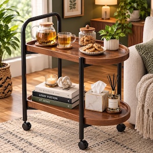 May include: A two-tiered rolling cart with a dark wood finish and black metal frame. The top shelf holds a glass teapot, a glass mug, a jar of cookies, and a small potted plant. The bottom shelf features stacked books, a candle, and a tissue box.