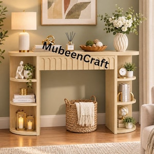 Puede incluir: Una mesa consola de madera clara con un panel frontal acanalado y estantes laterales curvos. La mesa presenta el texto MubeenCraft impreso en el centro. Está decorada con una lámpara blanca, libros, plantas, linternas y una cesta tejida.