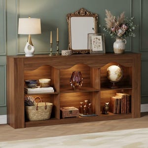 May include: A wooden console table with six open storage compartments featuring integrated interior lighting. The unit has a scalloped top edge and contains various decorative items including bowls, a glass vase, a globe, books, and a woven basket.