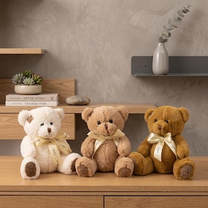 May include: Three plush teddy bears in cream, light brown, and dark brown colors, each sitting upright. Every bear features a cream-colored ribbon tied around its neck with the word Love printed on it and a patch on its foot reading I Love You.