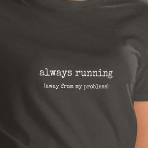 Może przedstawiać: Czarny t-shirt z krótkim rękawem z białym napisem w stylu maszyny do pisania wyśrodkowanym na klatce piersiowej o treści always running (away from my problems). Odzież posiada klasyczny okrągły dekolt oraz standardowy krój z gładkiej tkaniny.