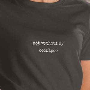 Puede incluir: Una camiseta negra de manga corta con el texto blanco "not without my cockapoo" impreso en una fuente estilo máquina de escribir en el centro del pecho. La prenda tiene un cuello redondo clásico y un diseño sencillo de color sólido.