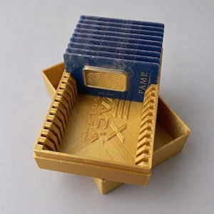 May include: A gold-colored 3D-printed storage tray with slotted sides, holding a row of blue PAMP Suisse gold bullion cards. The tray features the embossed text "VAULT" on its base. The cards are arranged vertically in the slots.