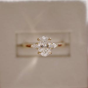 May include: A gold ring featuring a large oval-cut center stone held by four prongs, flanked by two smaller pear-cut stones on each side. The band is thin and polished, showcasing a classic three-stone design with clear, faceted gemstones.