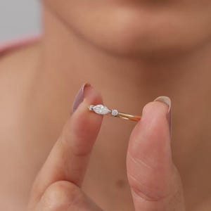 May include: A gold ring featuring a central marquise-cut diamond flanked by two smaller round-cut diamonds. The band is thin and polished, showcasing a delicate design with three clear stones set in a horizontal arrangement across the finger.