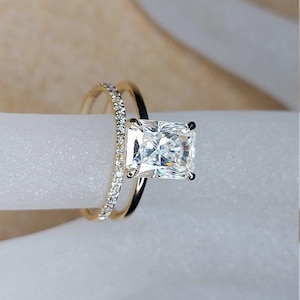 May include: A gold ring set featuring a large rectangular radiant-cut center stone held by four prongs. The set includes a plain gold band and a second thinner band encrusted with small round white stones along the top half of the shank.