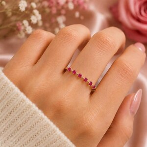 Könnte beinhalten: Ein goldener Ring mit einer Reihe von sieben kleinen, runden, leuchtend pinkfarbenen Edelsteinen, die entlang der Oberseite des dünnen, polierten Metallrings eingefasst sind. Der Ring wird an einem Finger getragen und zeigt das feine Design.