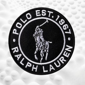 May include: A circular black embroidered patch featuring a white stitched border and the white text POLO EST. 1967 RALPH LAUREN arranged in a circle. In the center, a white embroidered graphic depicts a person riding a horse while holding a mallet.