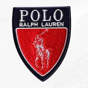 May include: A shield-shaped embroidered patch featuring a black border and a red center. The text POLO RALPH LAUREN is stitched in white at the top. A white embroidered graphic of a polo player on a horse is centered within the red section.