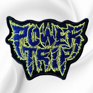 May include: An embroidered patch featuring the text Power Trip in a stylized, jagged font. The lettering is deep blue with a bright yellow outline, set against a black background with a distinct, irregular, wing-like shape and a thick black border.