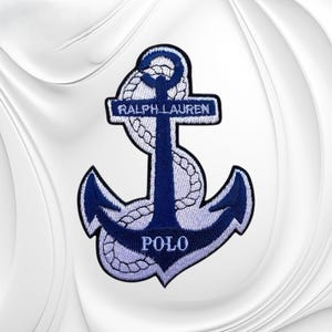May include: A navy blue and light blue embroidered patch featuring an anchor design wrapped in a rope. The text RALPH LAUREN is stitched across the horizontal bar of the anchor, and the word POLO is stitched in light blue on the lower section.