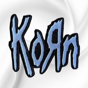 May include: A black embroidered patch featuring the band name Korn in a stylized, jagged light blue font. The lettering has a distinct, rough-edged appearance with a thick black border surrounding the light blue thread work on the logo design.