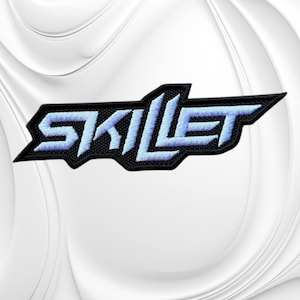 May include: An embroidered patch featuring the word SKILLET in a stylized, angular font. The lettering is light blue with a thick black border, creating a sharp, geometric design. The embroidery texture is visible throughout the entire logo.