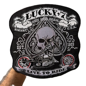 May include: A black embroidered patch featuring a central skull inside a spade shape, a motorcycle, and the text LUCKY 7, BIKES BOOZE BROADS, BIKER, U.S.A., EST. 1903, and LIVE TO RIDE. The motorcycle wheels have red accents and white spokes.