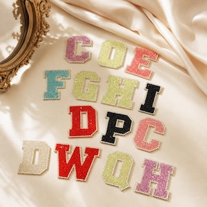 May include: A collection of varsity-style letter patches featuring a sparkling rhinestone finish. The letters include C, O, E, F, G, H, I, D, P, W, Q, and H in various colours such as pink, yellow, red, blue, black, and white, arranged on a fabric surface.