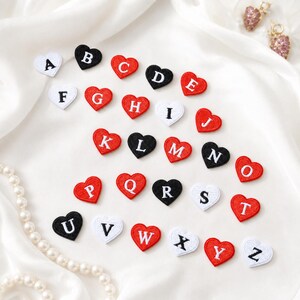 Puede incluir: Una colección de veintiséis parches bordados en forma de corazón, cada uno con una letra del alfabeto de la A a la Z. Los parches alternan entre corazones rojos con letras blancas y corazones negros o blancos con letras contrastantes.