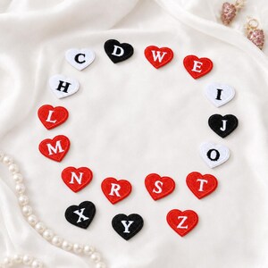 Op de afbeelding: Een verzameling hartvormige geborduurde patches gerangschikt in een cirkel. Elke patch bevat een enkele letter in een schreeflettertype. De harten zijn rood, wit of zwart, met contrasterende letters in wit of zwart garen.