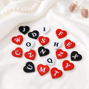 May include: A collection of heart-shaped embroidered patches in red, black, and white. Each patch features a single capital letter stitched in a contrasting color. The letters include J, D, I, F, W, O, S, H, E, C, Z, N, Q, X, U, M, T, and Y, arranged in a scattered pattern.