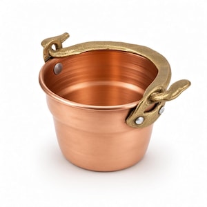 May include: A small, round copper bucket with a polished metallic finish. It features a textured, antique-gold colored metal handle attached to the sides with visible silver-toned rivets. The interior is smooth and reflects light, showing a consistent copper tone throughout the vessel.