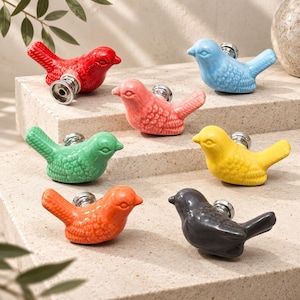 Puede incluir: Un conjunto de seis pomos para armarios con forma de pájaro en cerámica, disponibles en rojo, azul claro, rosa, verde, amarillo, naranja y gris oscuro. Cada pomo presenta un detalle de ala texturizada y una base metálica plateada.