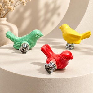 May include: Three bird-shaped cabinet knobs in bright red, yellow, and light green ceramic. Each knob features a glossy finish, detailed wing and tail textures, and a circular silver-toned metal base with a central screw hole for mounting.