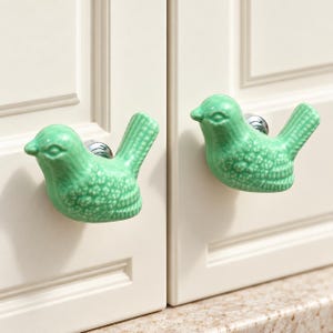 May include: Two mint green ceramic bird-shaped cabinet knobs with a glossy finish and textured wing details. Each bird figure is attached to a circular silver-toned metal base, designed for mounting onto cabinet doors or drawer fronts.