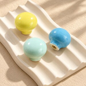 May include: Three round ceramic cabinet knobs in bright yellow, light blue, and pale mint green. Each knob features a smooth, glossy finish and a central metal threaded insert on the base for mounting, displayed on a white wavy ceramic tray.