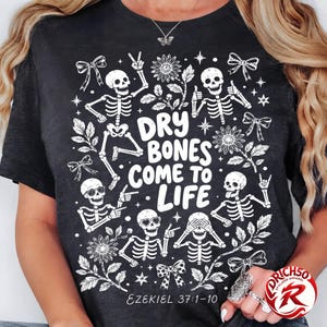 May include: A black t-shirt featuring a white graphic print of five skeletons in various poses, surrounded by flowers, bows, and stars. The text reads "DRY BONES COME TO LIFE" in the center, with "EZEKIEL 37:1-10" printed in smaller white text at the bottom.