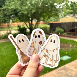 Op de afbeelding: Drie gestanste vinylstickers met cartoongeesten. Eén geest houdt een bruine bezem vast, een andere heeft een patchworkpatroon met oranje en bruine stoffen vierkantjes, en de derde is gedrapeerd met een lichtsnoer van oranje stervormige lampjes.