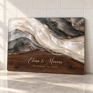 May include: A rectangular cutting board featuring a dark wood grain base with the white printed text Elena & Marcus and OCTOBER 10, 2027. The upper portion displays an abstract marbled design in shades of cream, grey, and black with gold accents.