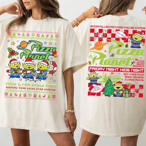 May include: An off-white t-shirt featuring Pizza Planet graphics. The front shows three green aliens in Santa hats with a candy cane and holiday patterns. The back displays Pizza Planet branding, aliens, a Christmas tree, and text including Interstellar Fast-Food Fueling Station and Friday Night Kids Night.