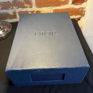 Peut inclure: Une boîte rectangulaire bleu marine avec une finition texturée rappelant le cuir. Le mot DIOR est embossé en lettres argentées au centre du couvercle. La boîte présente une découpe rectangulaire sur le devant et une structure rigide.