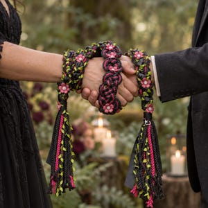Könnte beinhalten: Ein Handfasting-Band mit geflochtenem schwarzem und pinkem Design, verziert mit kleinen pinken Blüten und weißen Zentren. Das Band hat lange Quasten mit grünen Blattakzenten und schwarzen Bändern, die um zwei ineinander verschränkte Hände liegen.