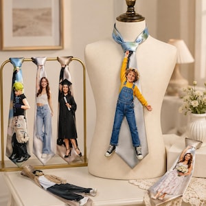Può includere: Una collezione di cravatte personalizzate con stampe fotografiche a figura intera di varie persone. Una cravatta è esposta su un manichino, mentre altre sono appese a un supporto dorato o appoggiate in piano, mostrando vari outfit come salopette, toghe e abiti.