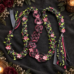 Puede incluir: Un cinturón o fajín trenzado decorativo que presenta cordón negro y magenta, entrelazado con cinta de satén negra y pequeñas hojas de tela verde. El accesorio está adornado con flores de tela rosa, cada una con un pequeño cristal transparente en el centro.