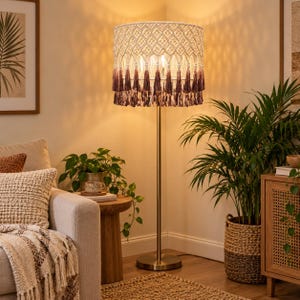 May include: A floor lamp with a cylindrical lampshade featuring cream-colored macrame knotting in a diamond pattern and a bottom fringe of dark purple tassels. The lamp has a slender metallic pole and a circular base, emitting a warm light.